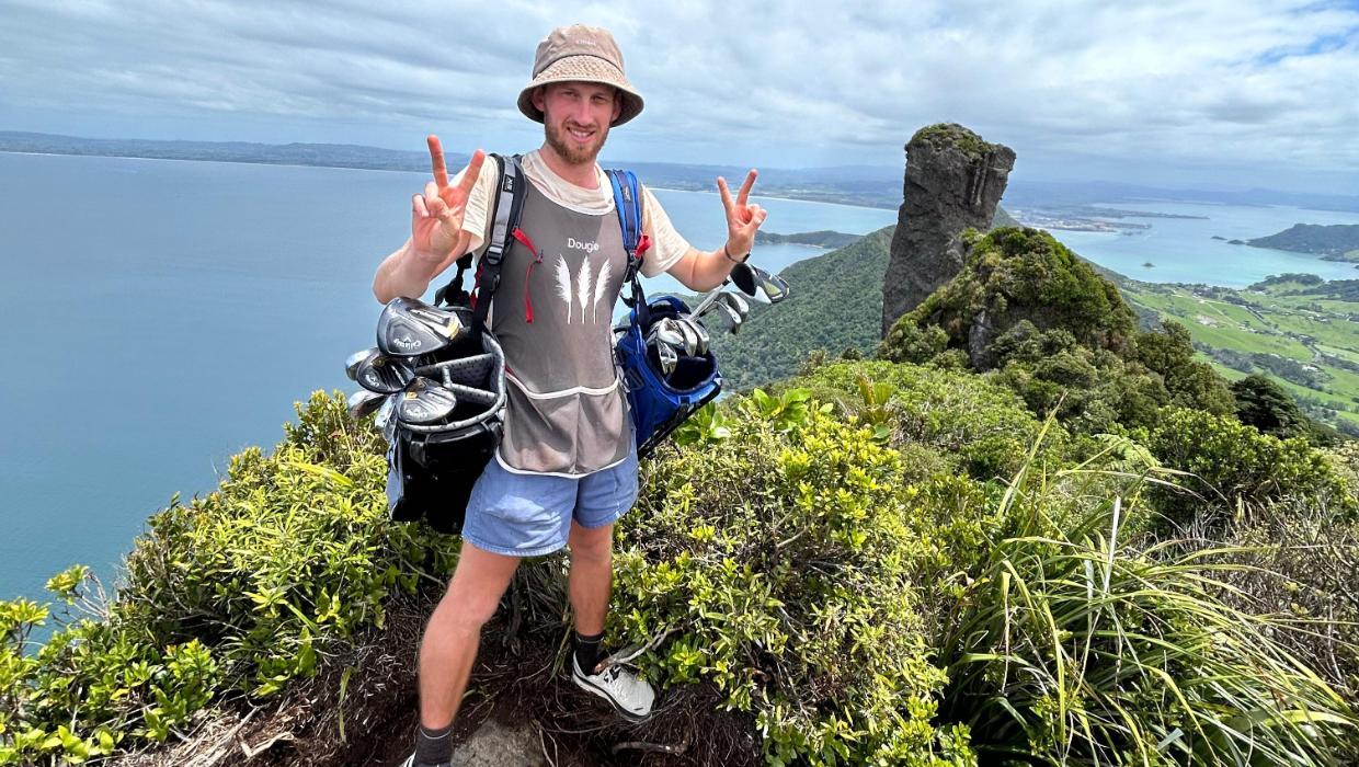 Man in a hat holding a golf bag on a lush hilltop, making peace signs, with ocean and coastline in the background under a cloudy sky