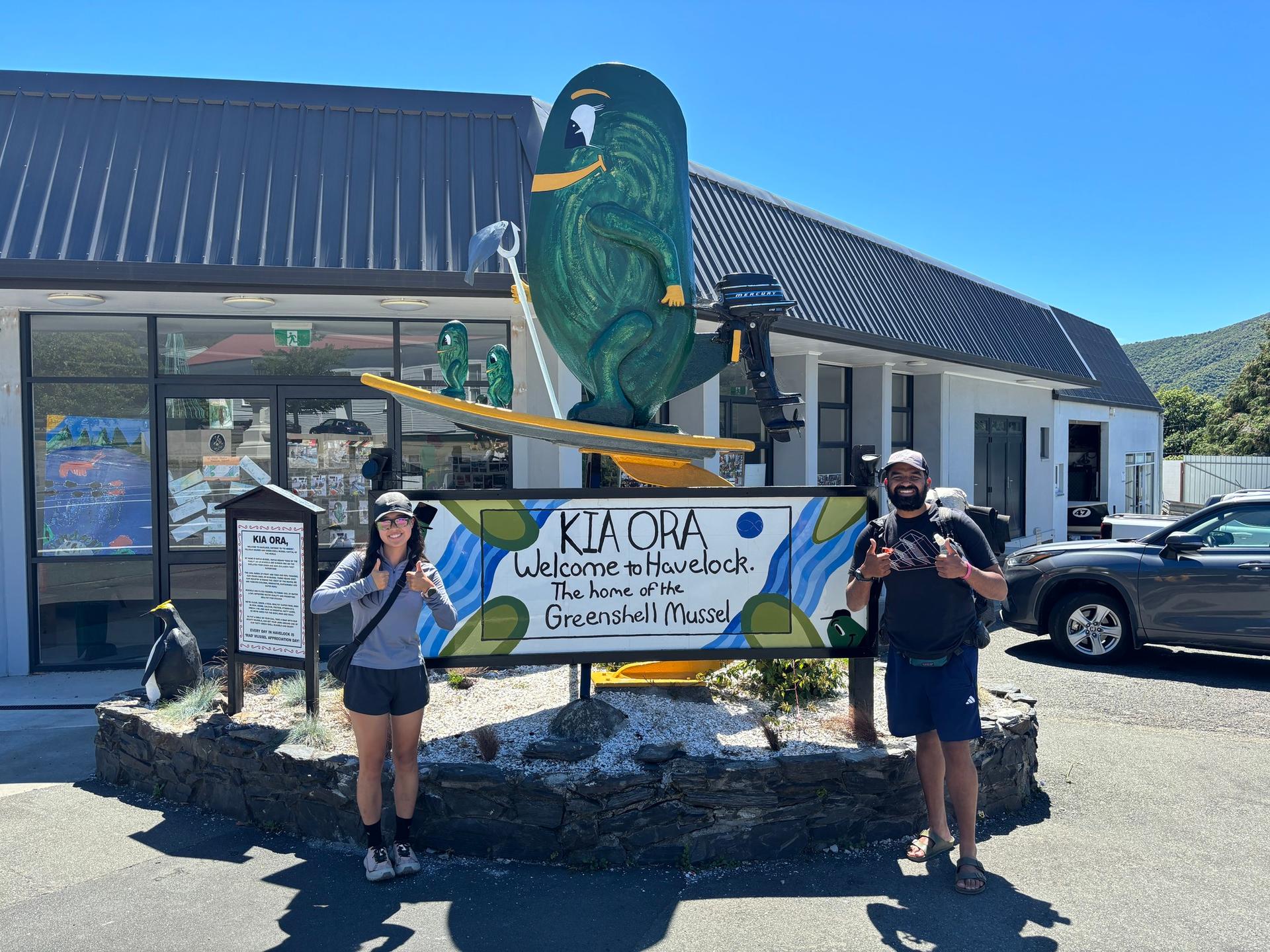Two people stand in front of a welcome sign for Havelock, adorned with a large mussel sculpture, on a sunny day