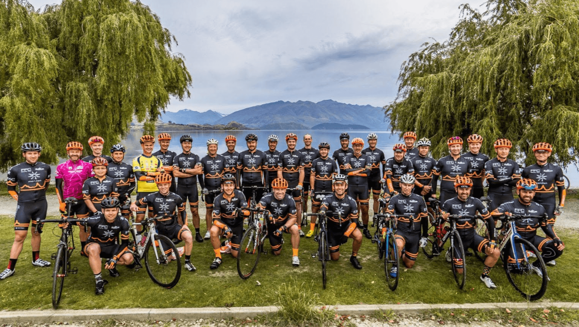 A large group of cyclists in matching jerseys and helmets pose with their bikes in front of a lake and mountains, flanked by trees