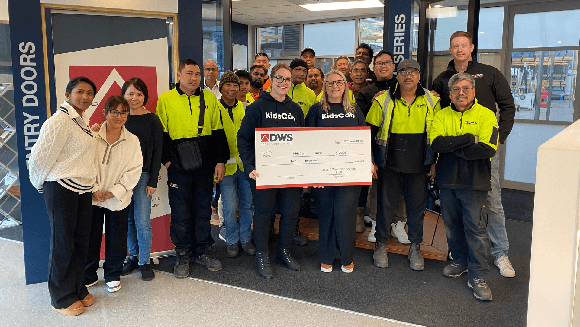 A group of people, some in bright safety jackets, stand indoors holding a large check. They're smiling and posing for the photo
