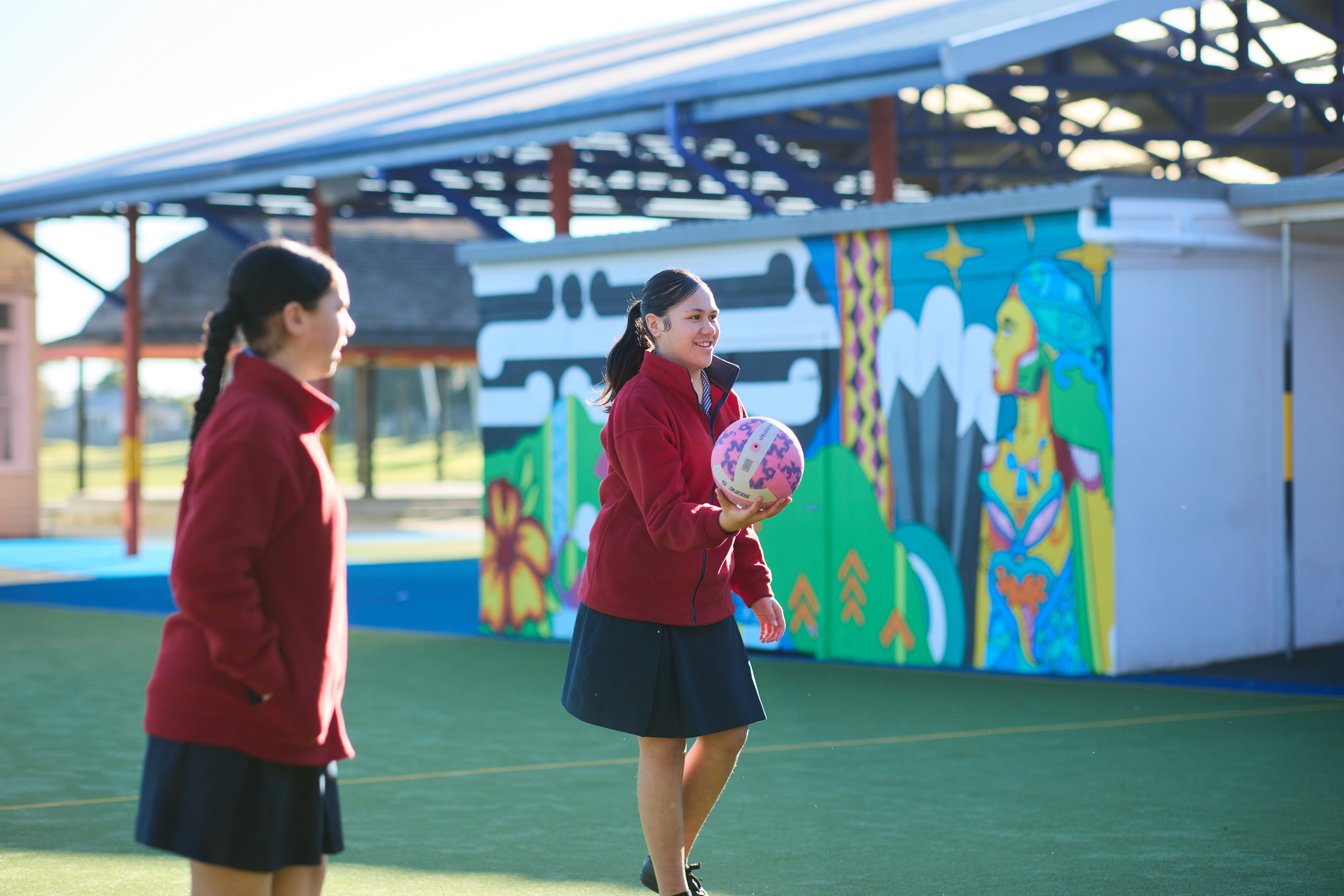 Two students in red jackets play with a colorful ball on a school court, with vibrant murals on the building behind them.